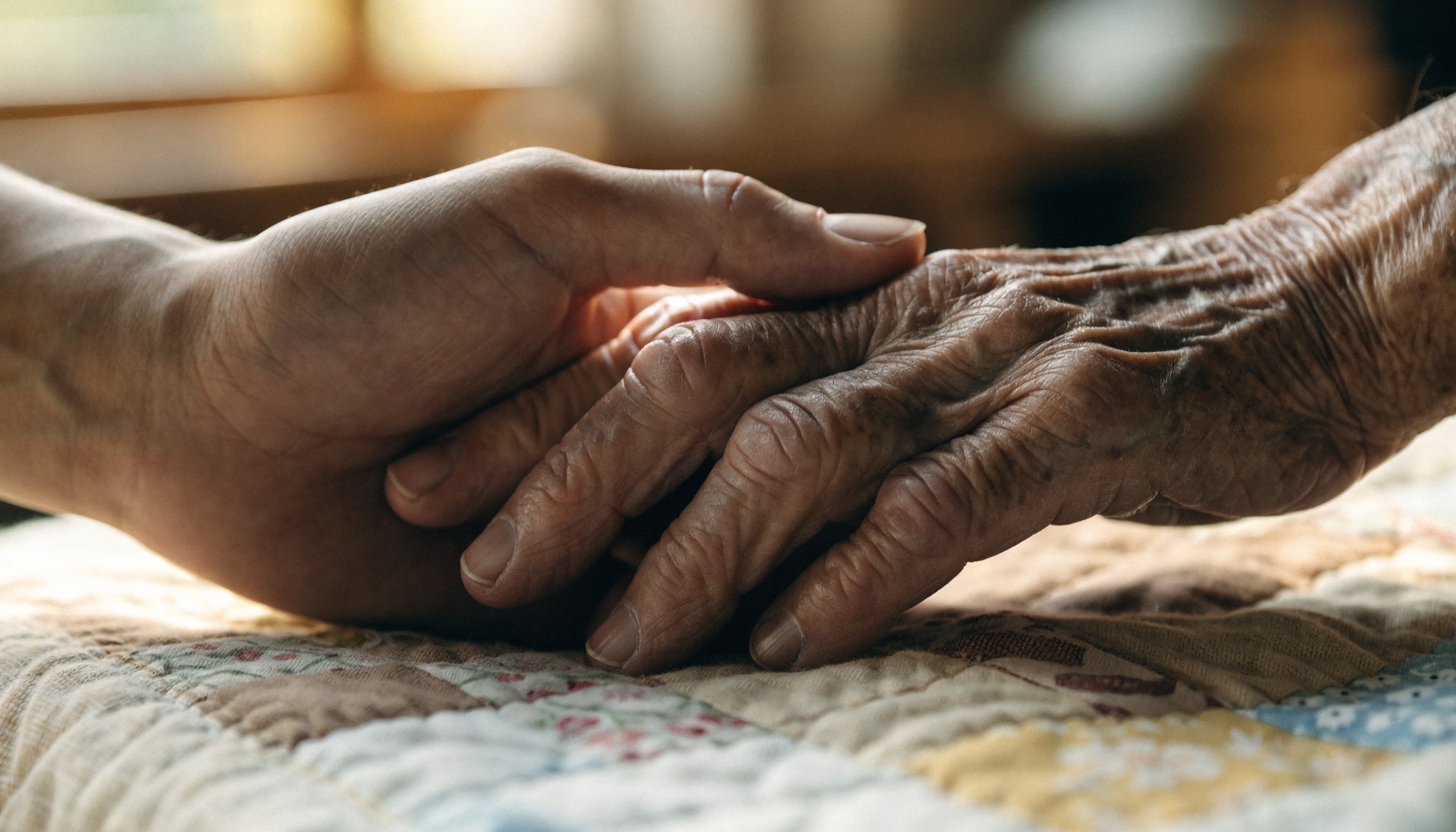 A caregiver's hand gently holding an elderly person's hand on a handmade quilt